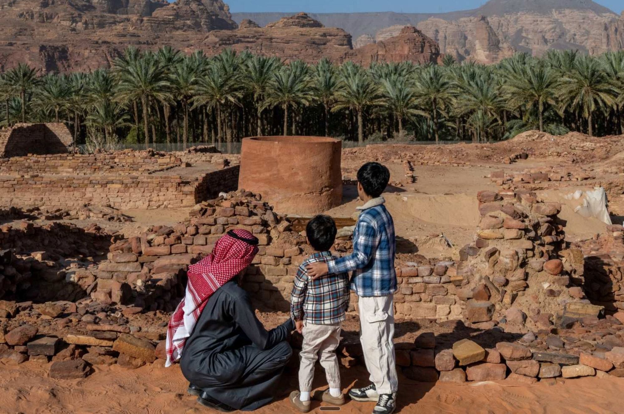 A family looking at stone ruins in front of a palm grove and red cliffs in AlUla