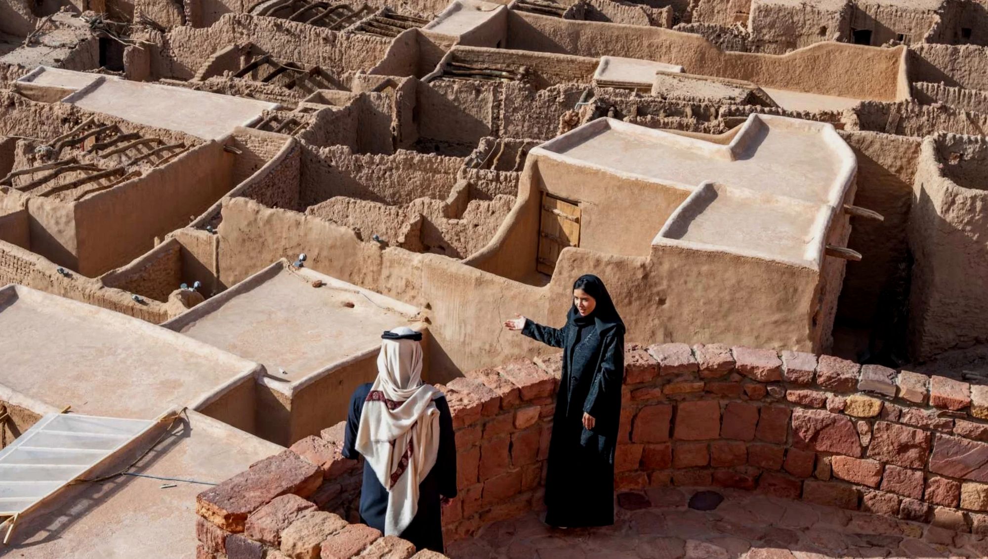 Two people in traditional dress standing among the mud-brick rooftops of AlUla’s old town
