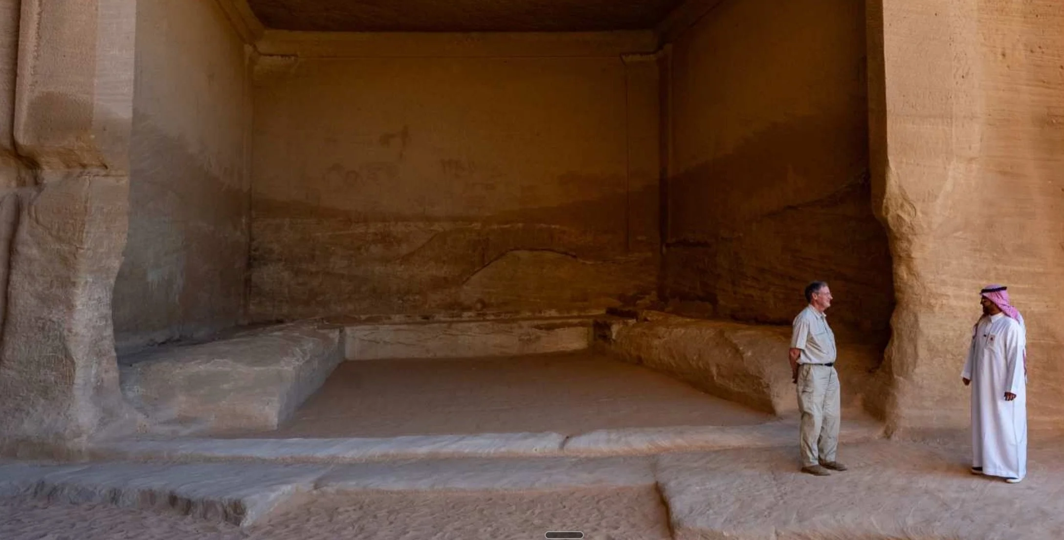 Two men standing inside a large chamber carved into a sandstone cliff in AlUla
